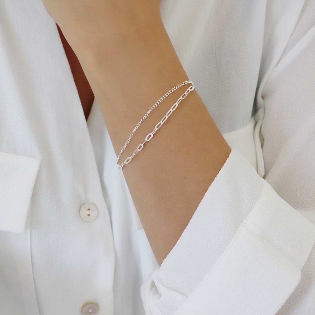 A delicate silver chain bracelet worn on a person's wrist, shown against a white button-down shirt.