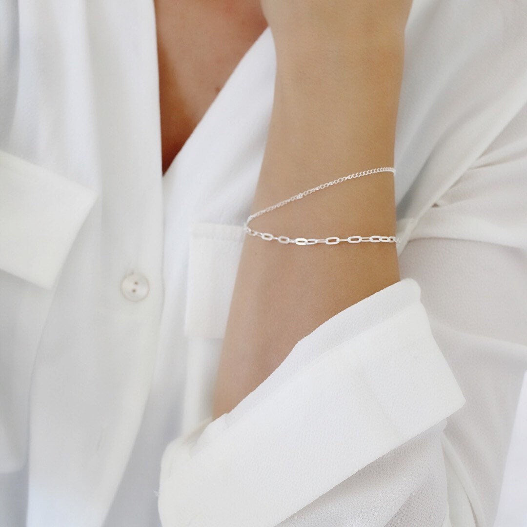 A delicate silver chain bracelet with open oval links worn on a person's wrist, shown against a white button-down shirt.