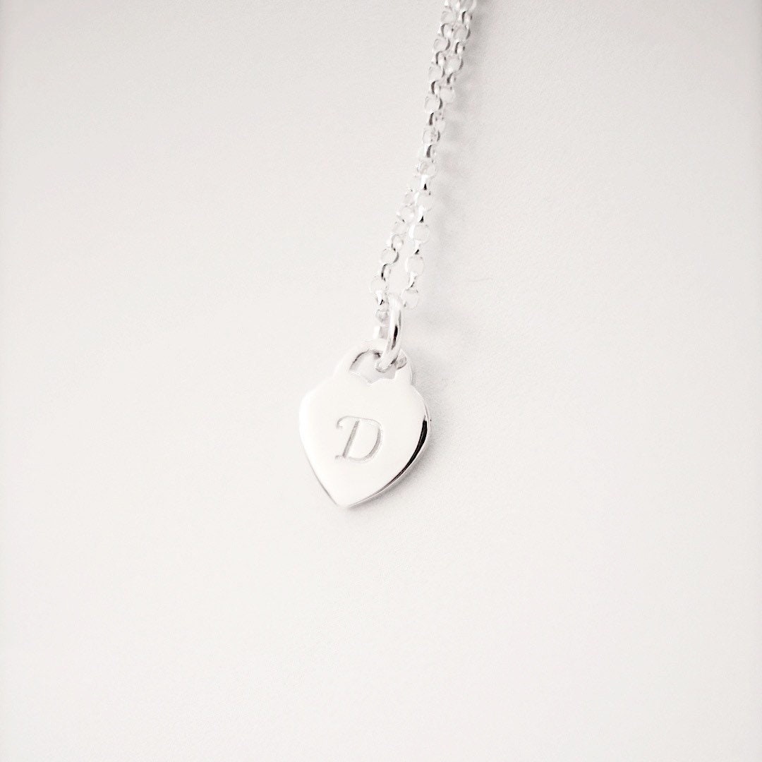 A silver heart-shaped pendant necklace with a single engraved letter 'D' on a delicate silver chain, set against a plain light gray background.