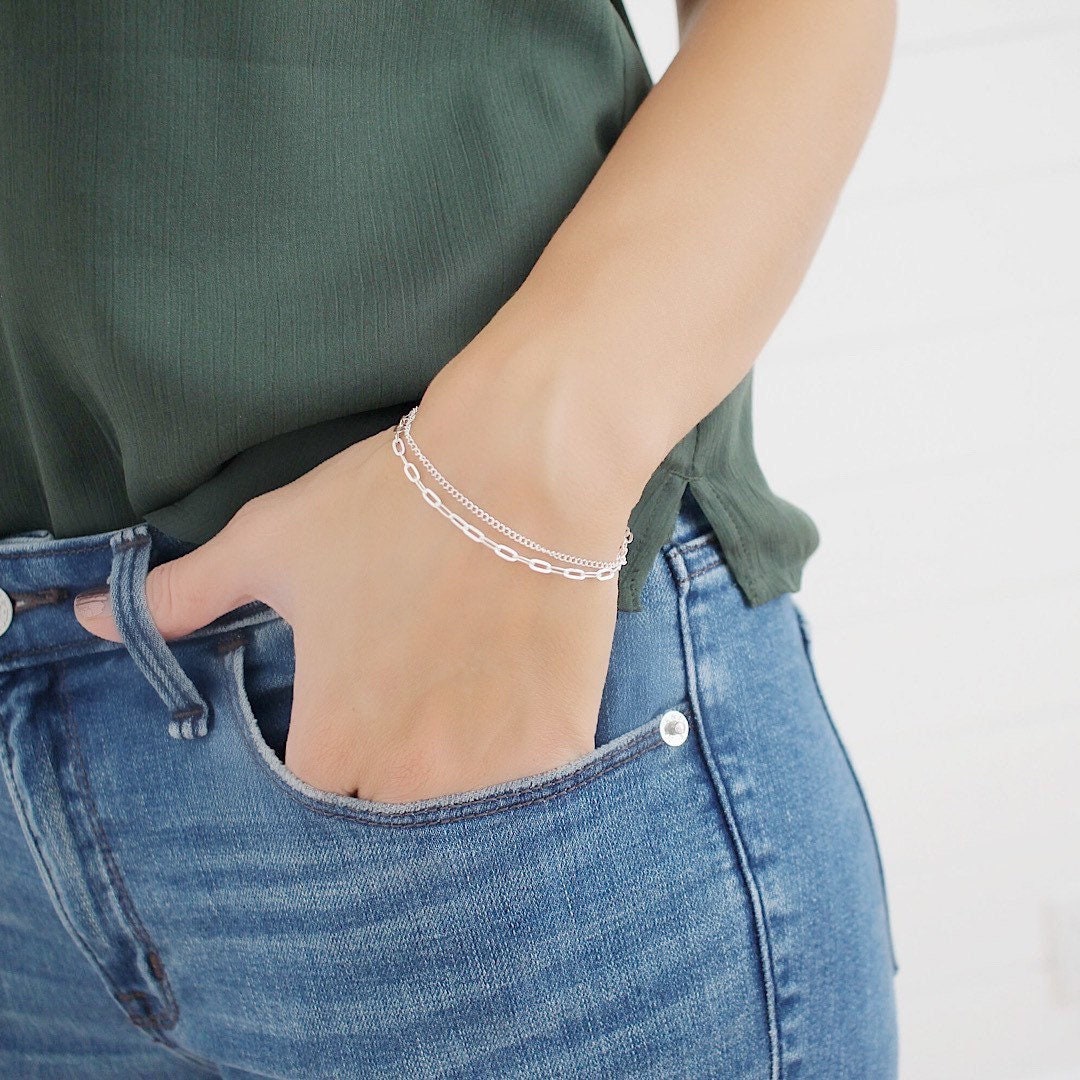 A close-up of a person wearing a delicate silver chain bracelet on their wrist, paired with a dark green top and blue jeans.
