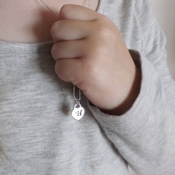 A close-up of a person wearing a silver necklace with a heart-shaped pendant featuring the letter 'H', while wearing a light gray long-sleeve top.