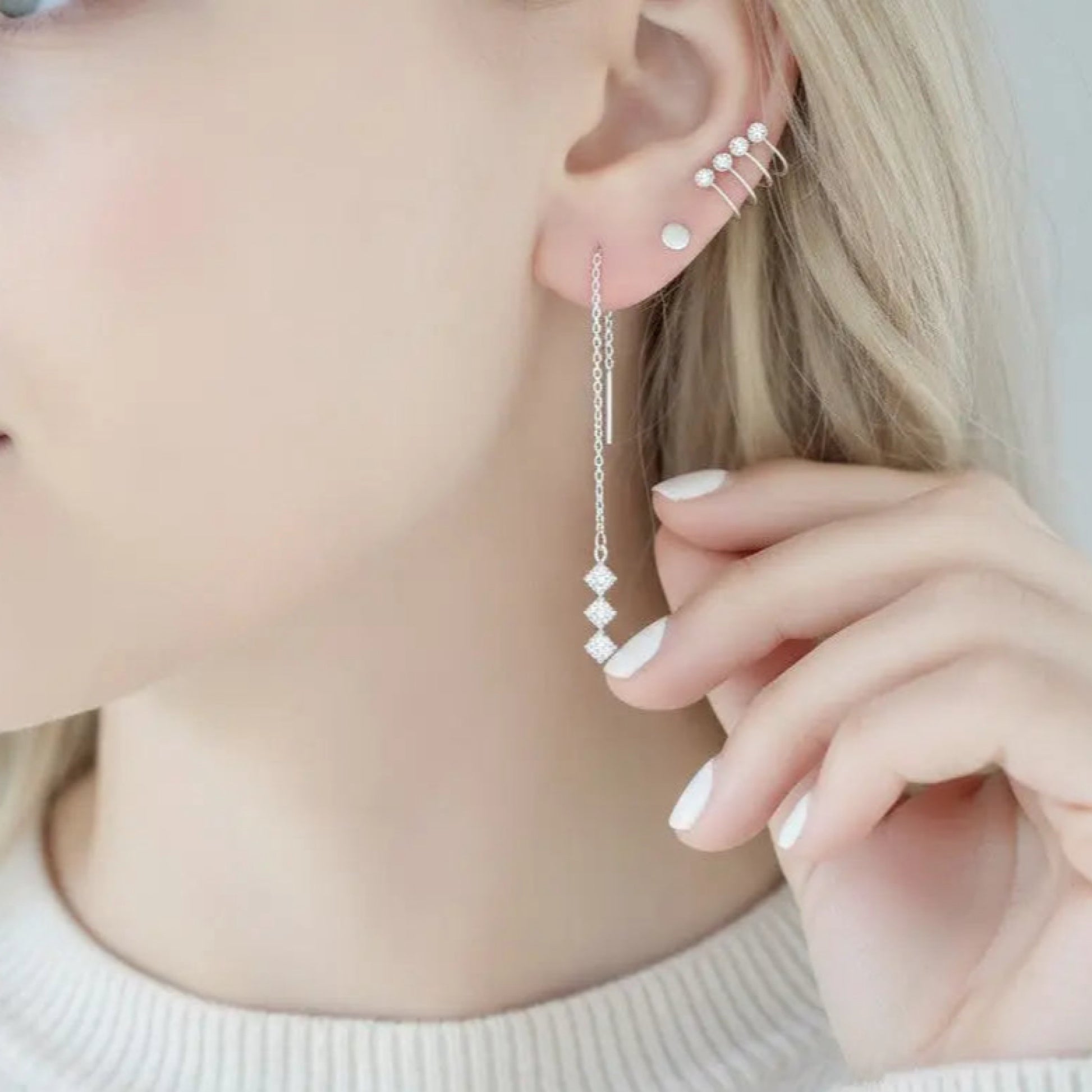 A close-up of a model's ear wearing a long silver dangle earring with small diamond-shaped beads, along with stacked ear cuffs.