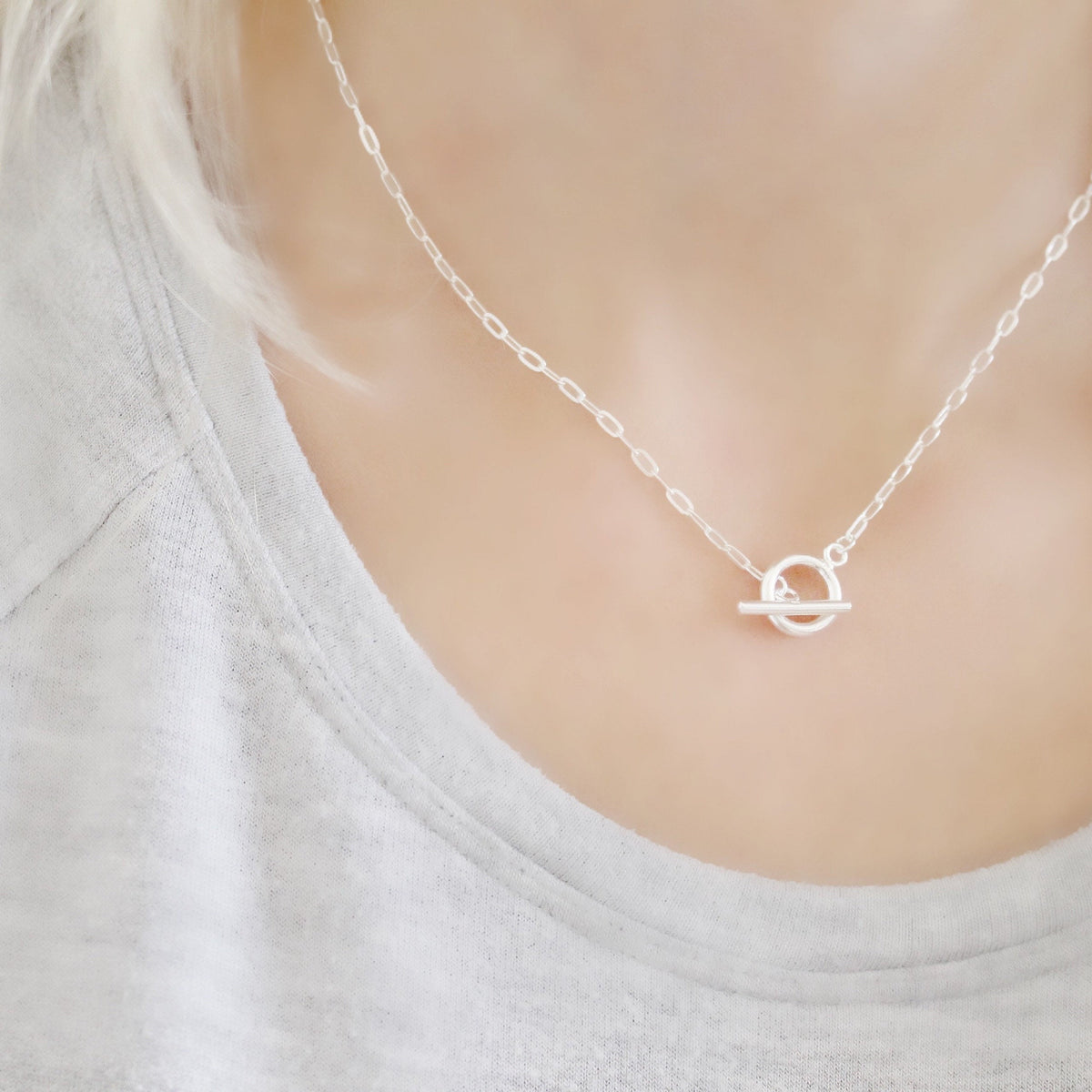 A close-up of a delicate silver chain necklace with a minimalist circular pendant, worn by a person in a light gray top.