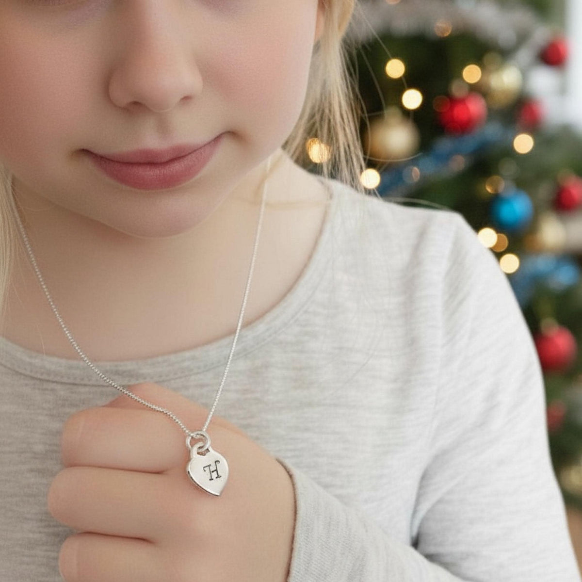 A close-up lifestyle shot of a child wearing a delicate silver chain necklace with a small heart-shaped pendant engraved with the letter 'M', set against a festive blurred Christmas tree background.