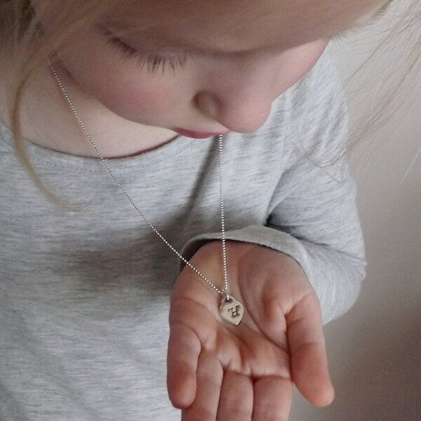A close-up lifestyle shot of a young child wearing a delicate silver chain necklace with a small heart-shaped pendant resting in their hand.