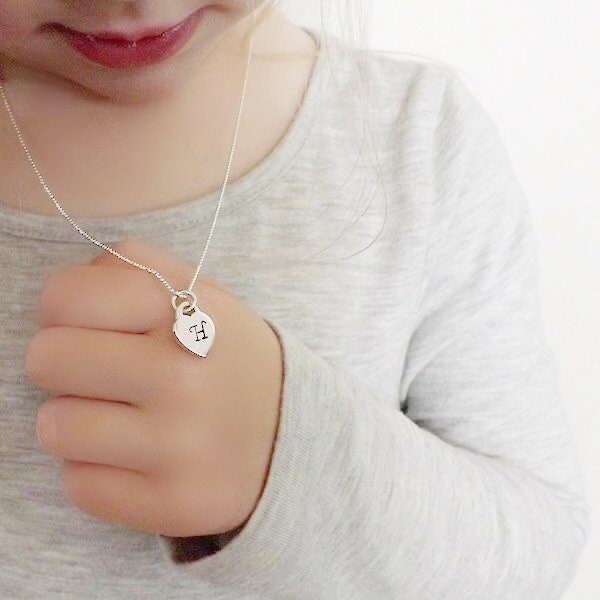 A close-up of a child wearing a delicate silver necklace with a small heart-shaped pendant engraved with a monogram, held gently by an adult hand against a light gray shirt.