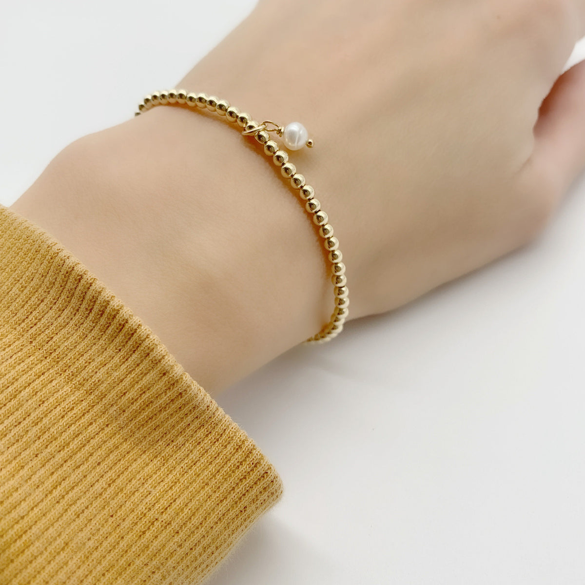 A delicate gold beaded bracelet with a small white pearl charm, shown worn on a person's wrist with a mustard yellow ribbed sweater cuff visible.